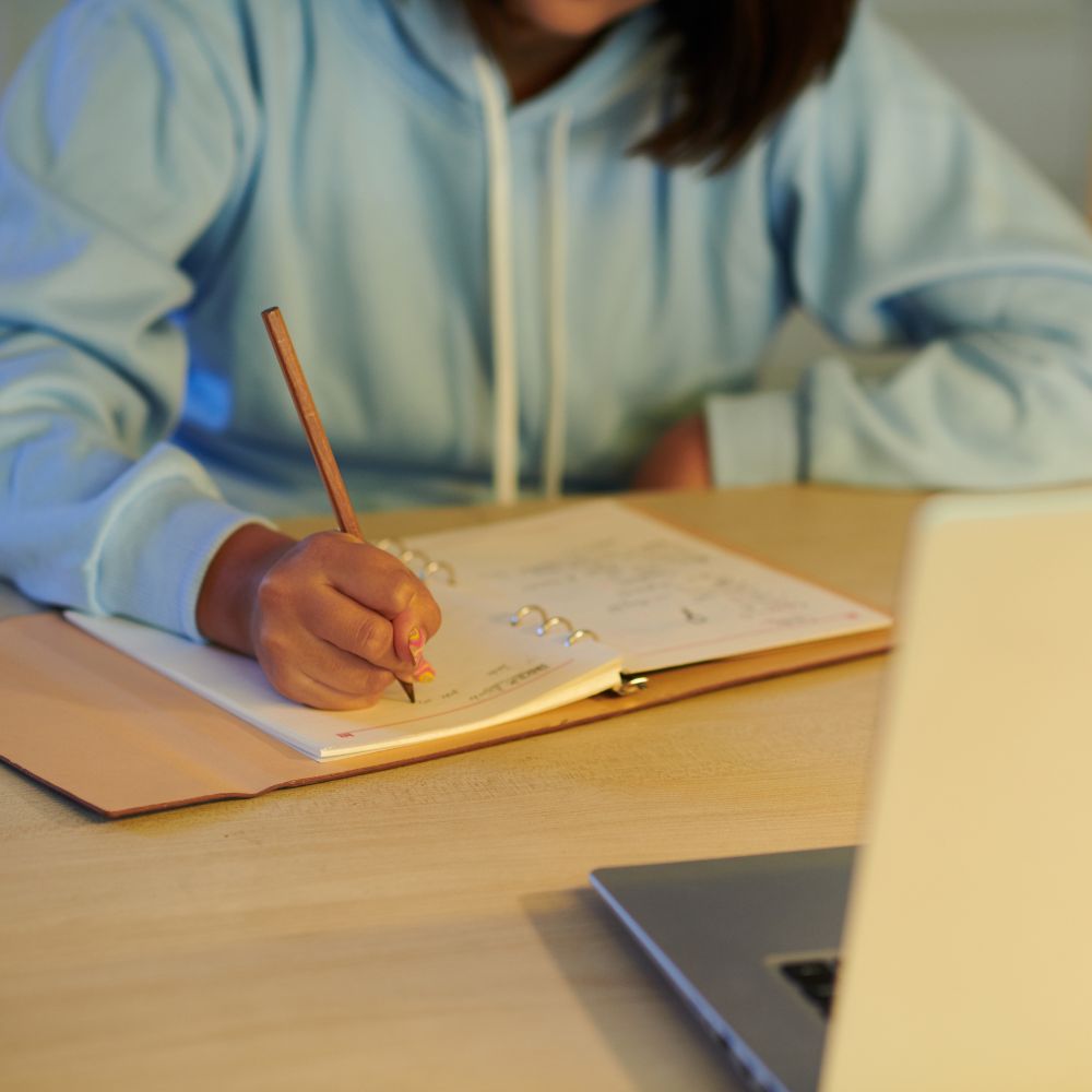 Student writing in journal beside open laptop screen