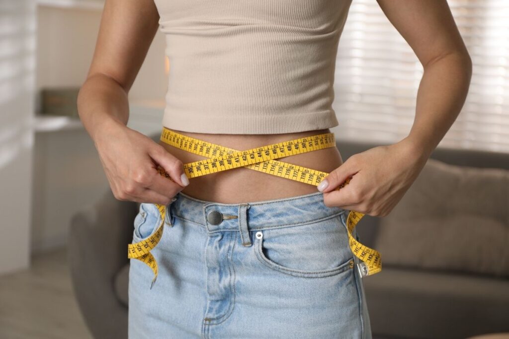 Woman in a crop top and jeans measuring her waist with a tape measure