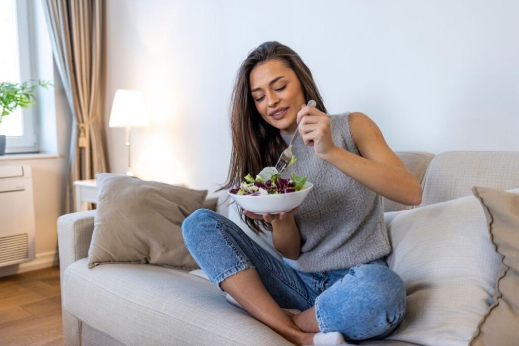 Woman sitting cross-legged on a couch, smiling while eating a fresh salad