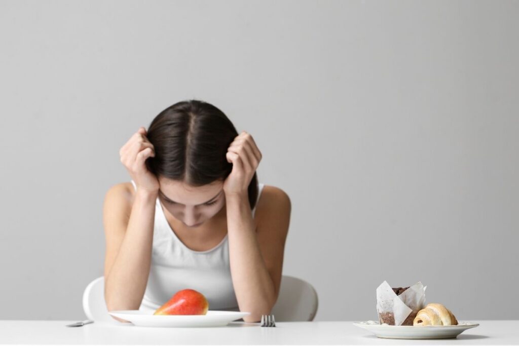 Young woman sitting at a table looking down and staring at a pear on her plate while a plate of desserts sits off to the side