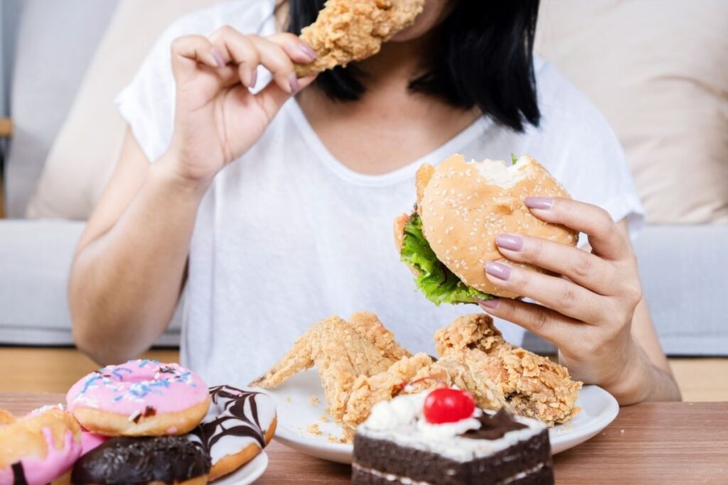 Woman engaging in binge eating, surrounded by fried foods, burgers, and desserts