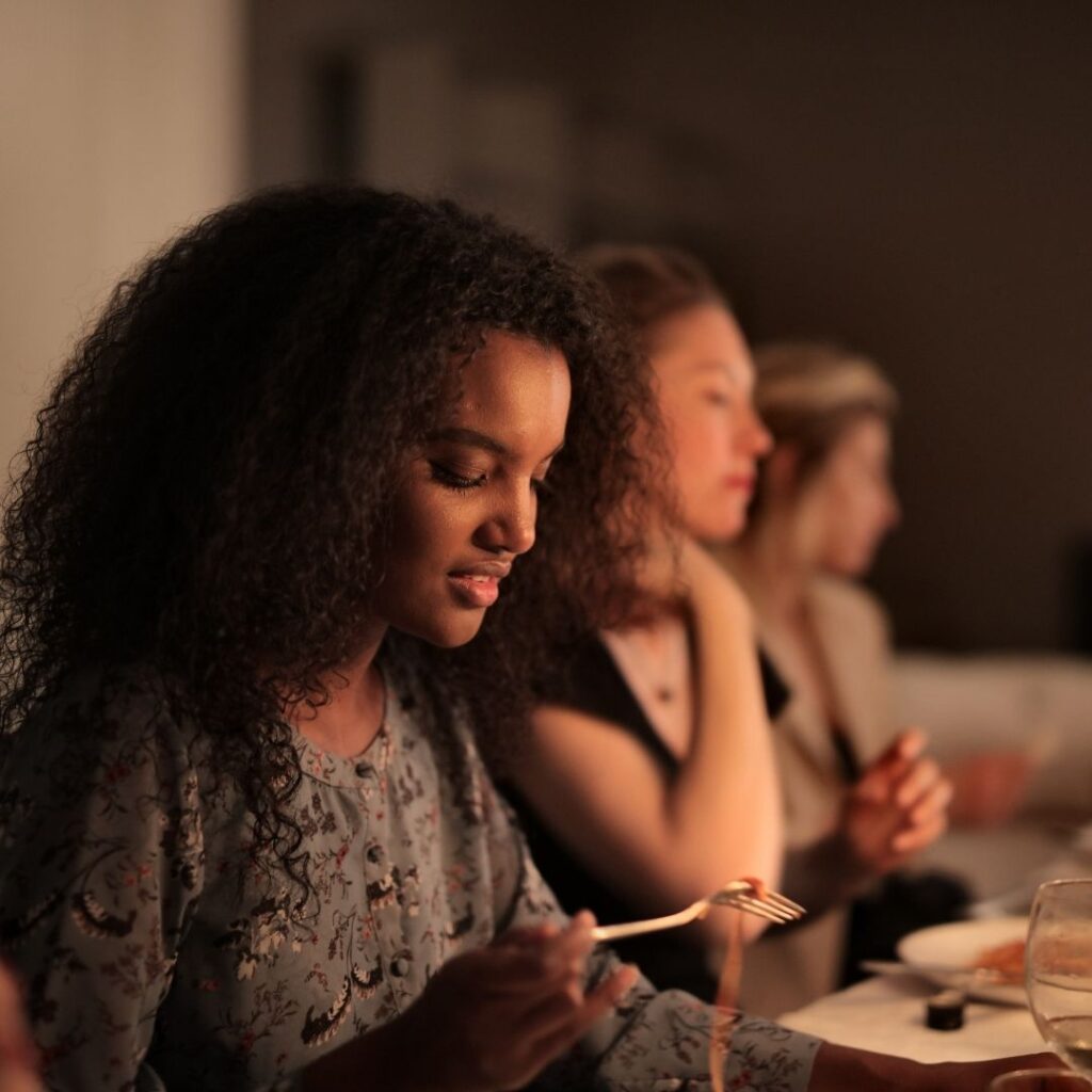 A woman with curly hair eats pasta from a fork, sitting at a table with other people 