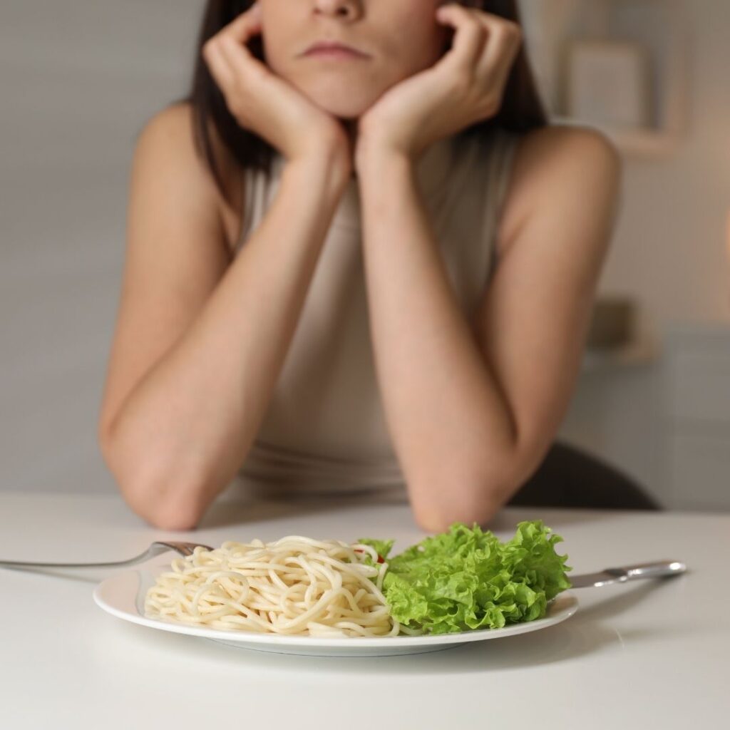 A woman looks sadly at a plate of spaghetti and lettuce, resting her chin in her hands