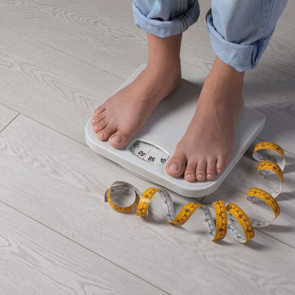 A person stands barefoot on a scale with a measuring tape coiled beside them