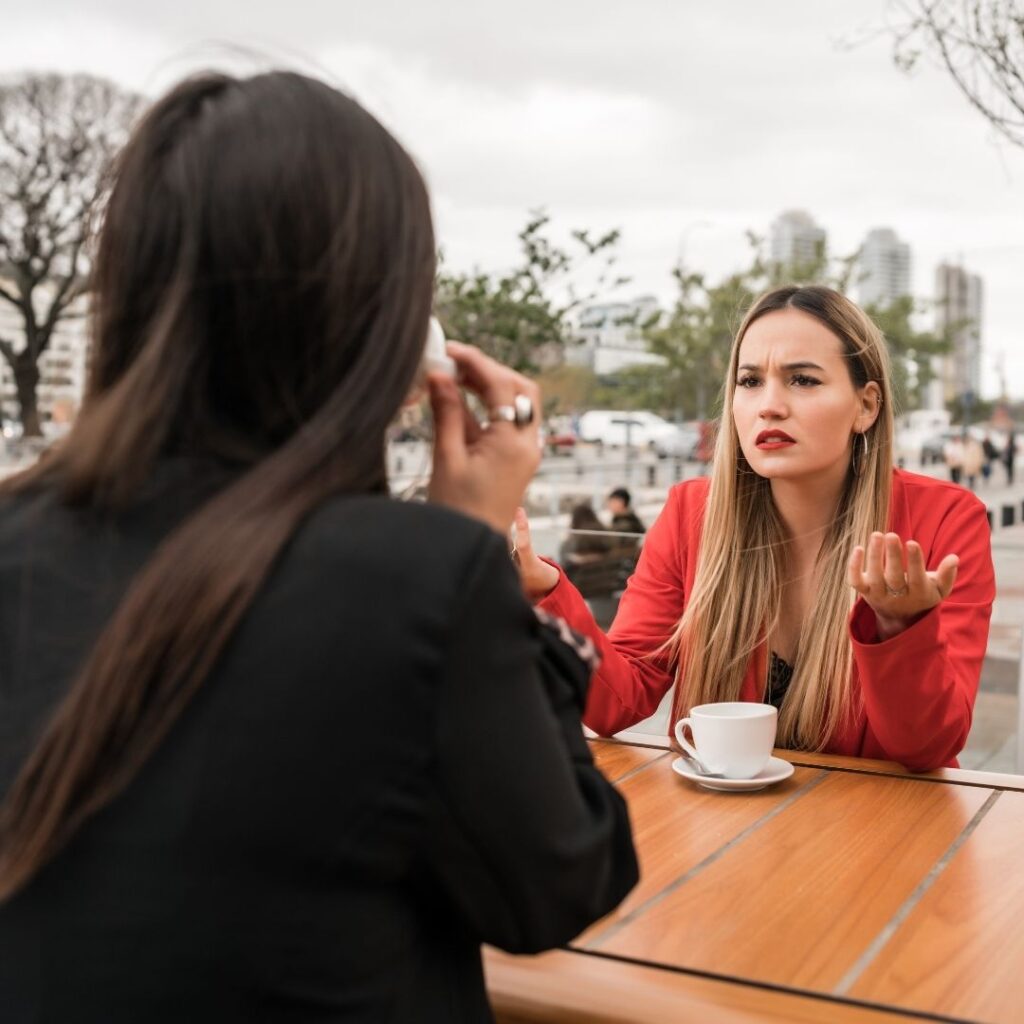 A woman in a red jacket gestures with frustration while talking to a friend sitting across at an outdoor café