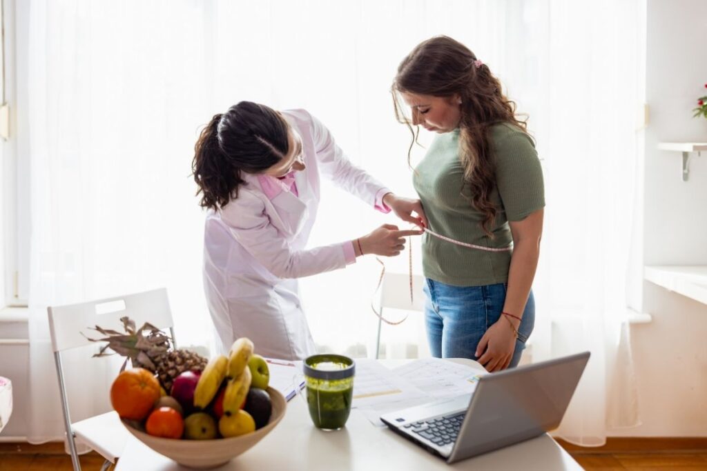 Healthcare professional measuring a woman’s waist with a tape measure in a brightly lit room, with a bowl of fruit and a laptop on the table
