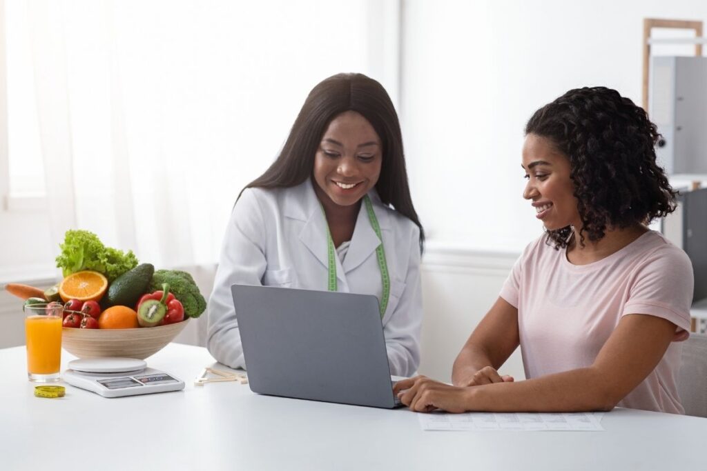 Nutrition therapist and client smiling while looking at a laptop during a consultation, with a bowl of fresh produce and a glass of juice on the table