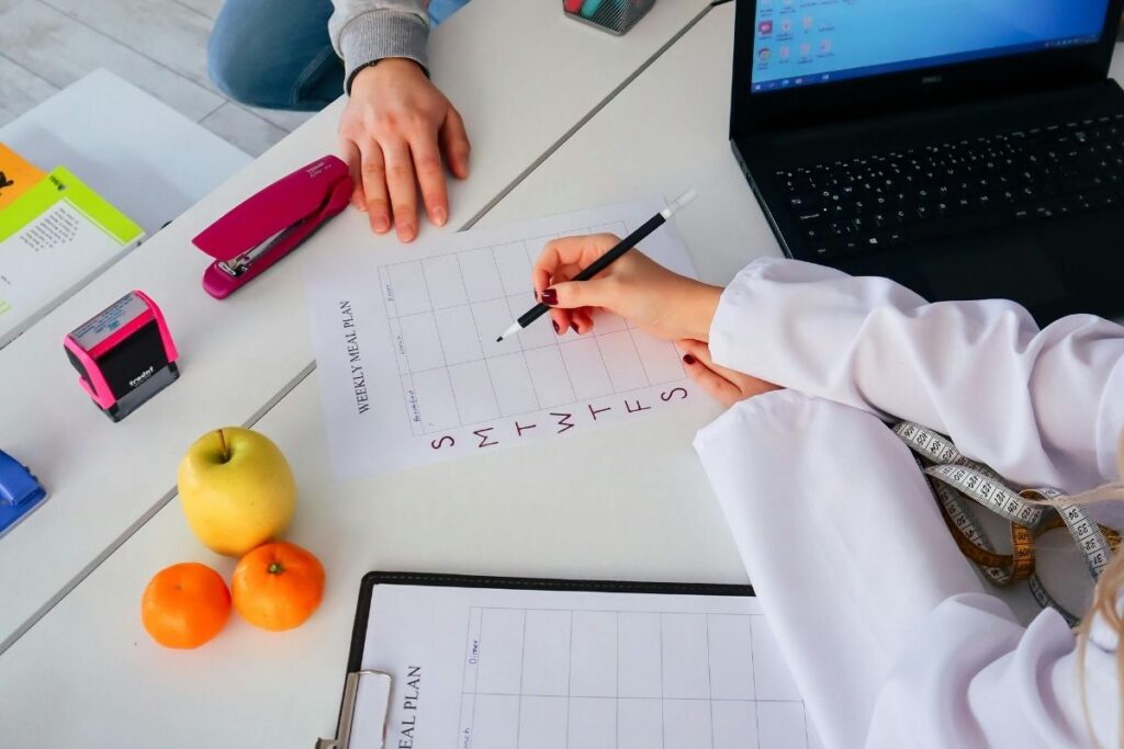 Close-up of hands filling out a weekly meal plan at a desk with fruit, stationery, and a laptop