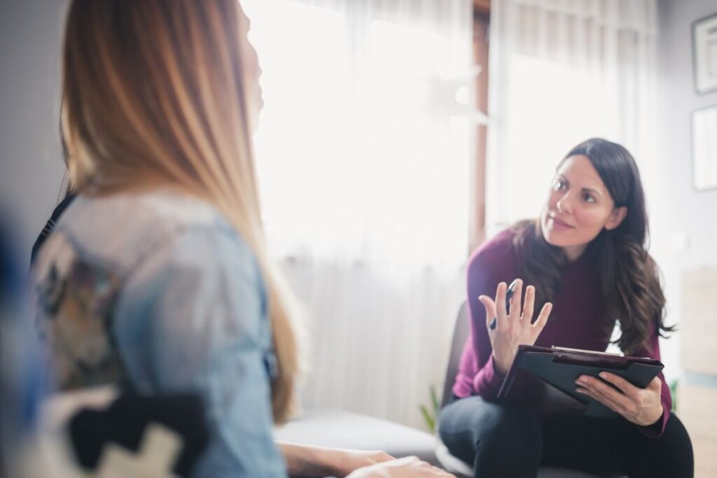 Therapist holding a clipboard and speaking with a client during a one-on-one session in a softly lit room
