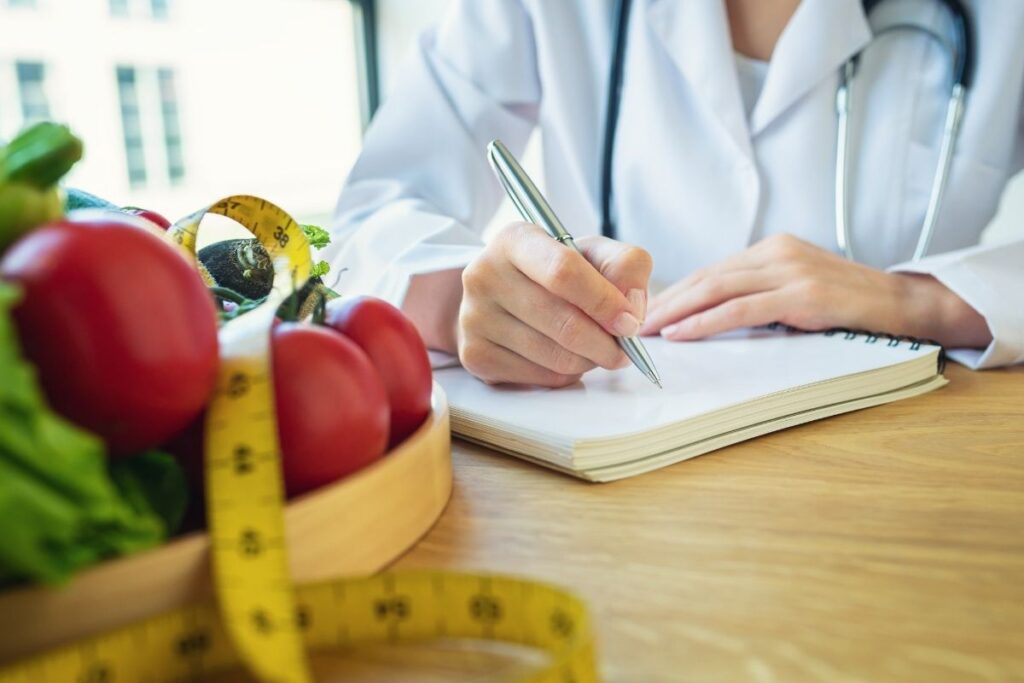Close-up of a healthcare provider writing in a notebook next to a tray of fresh vegetables and a yellow measuring tape
