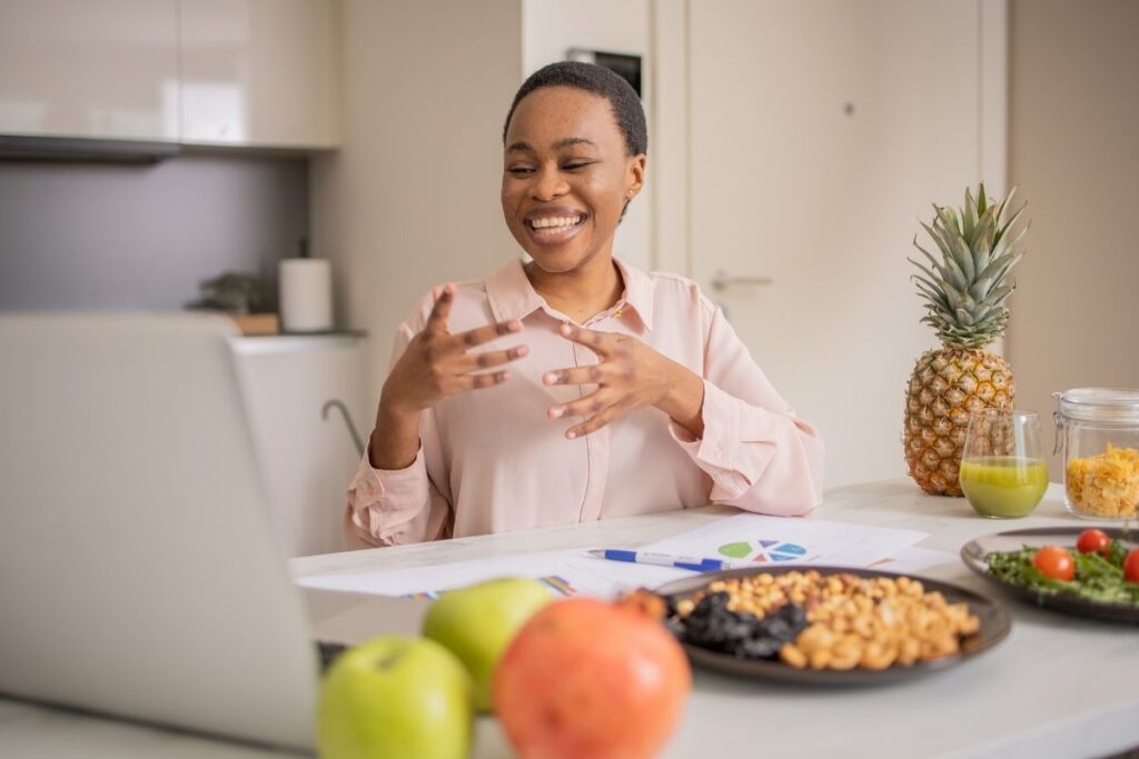 Smiling woman gesturing while speaking during a virtual consultation at a kitchen table with charts, fruit, and a laptop