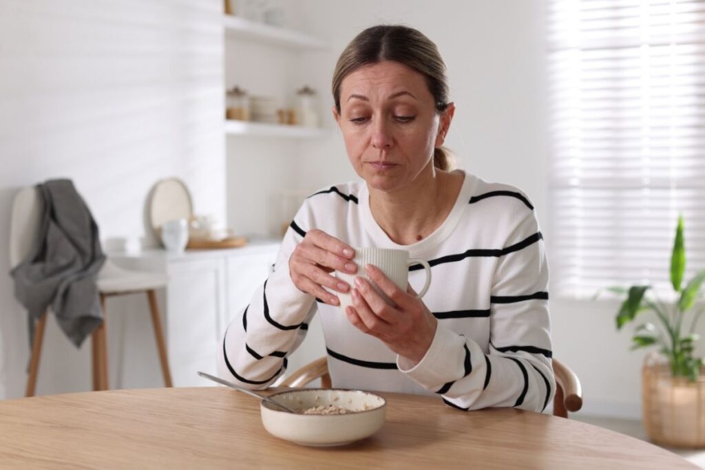 Woman sitting at a kitchen table holding a mug and looking thoughtful, with a bowl of cereal in front of her