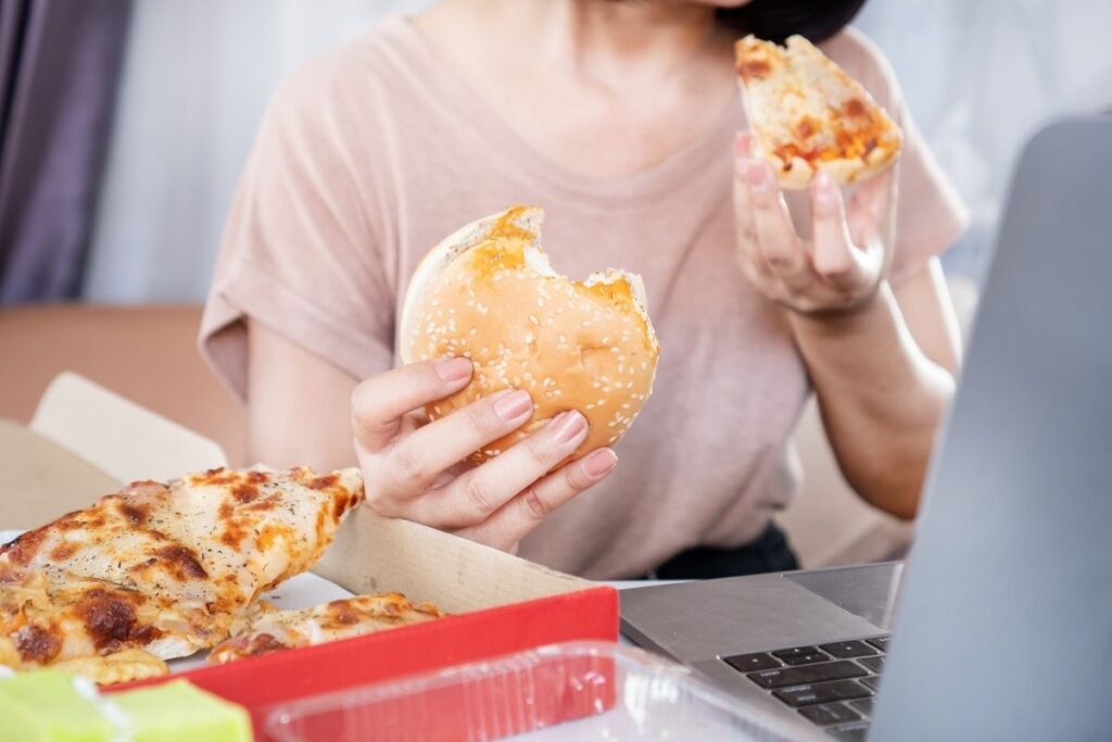 Woman holding a burger and pizza slice while eating in front of a laptop