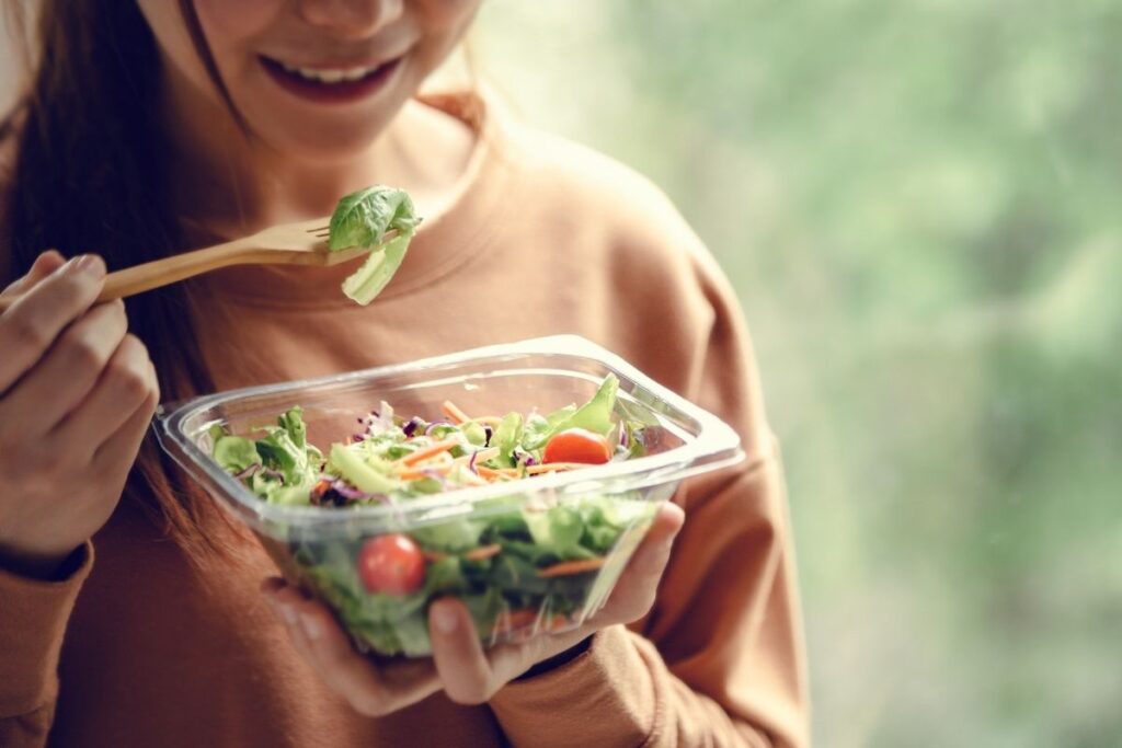 Close-up of a smiling woman eating a colorful salad from a plastic container with a wooden fork