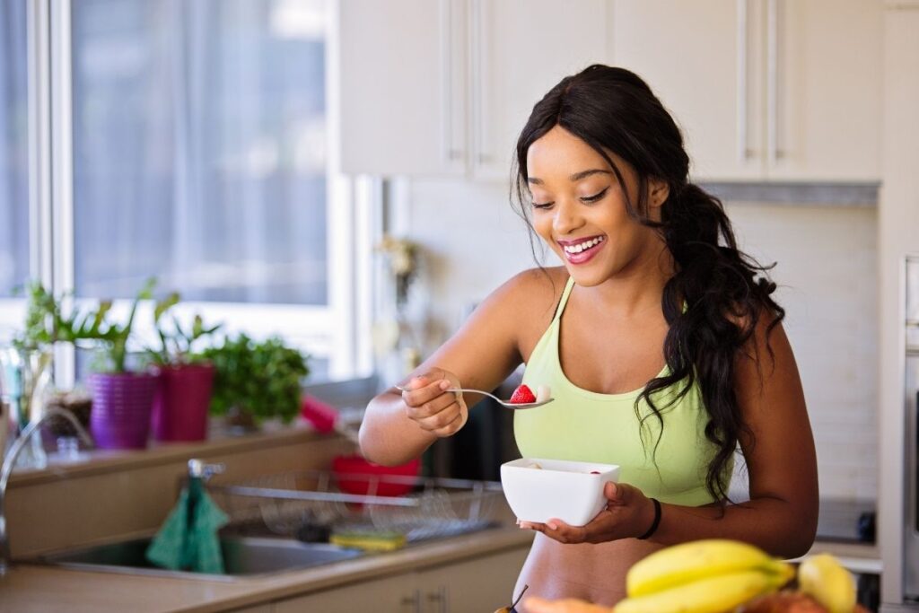 Smiling woman in athletic wear eating fruit from a bowl in a bright kitchen