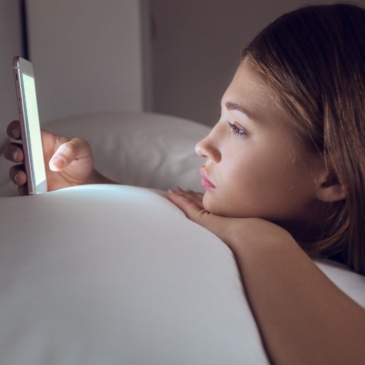Girl lying in bed at night, staring at a glowing phone screen with a thoughtful, serious look