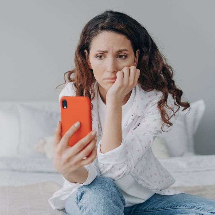 Worried woman sitting on bed, holding phone in one hand while resting her face on the other