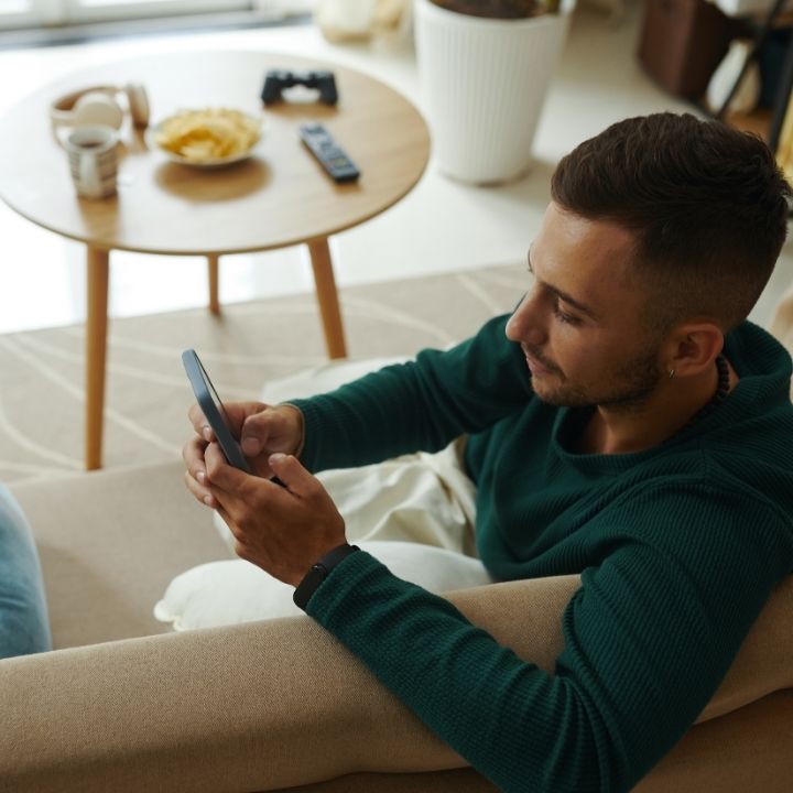 Man sitting on couch, holding and looking at phone while a table with snacks sits nearby