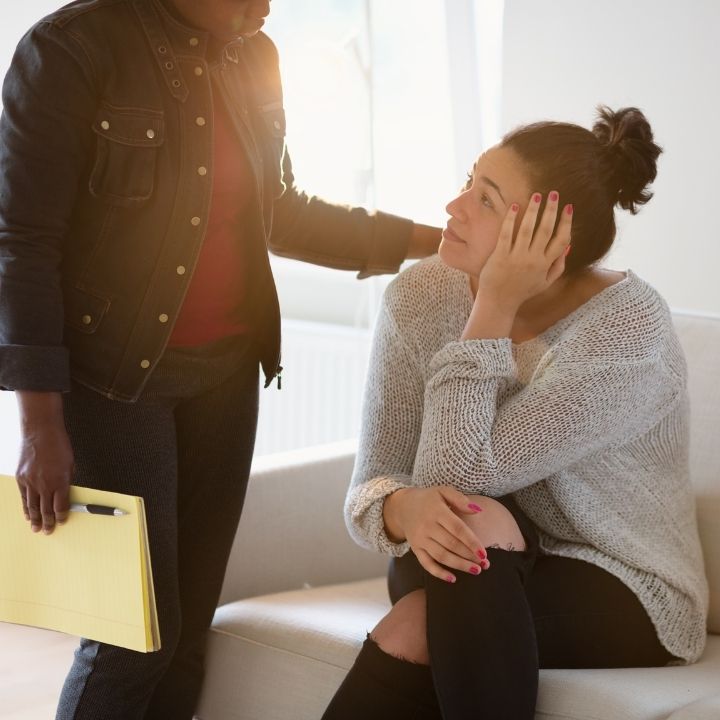 Woman sitting on couch, looking up emotionally while therapist offers comfort with a hand